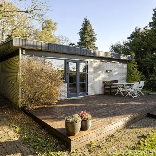 En træterrasse med to potteplanter står foran et hvidt murstenshus. Der er et bord og stole på terrassen. Haven er omkranset af høje træer og buske.