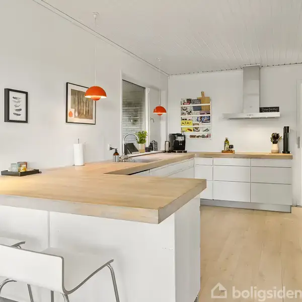 Et lyst, moderne køkken med hvide skabe og træbordplader. To hvide barstole står ved en ø. Orange pendellamper hænger over bordet, mens en plante ses i vinduet.