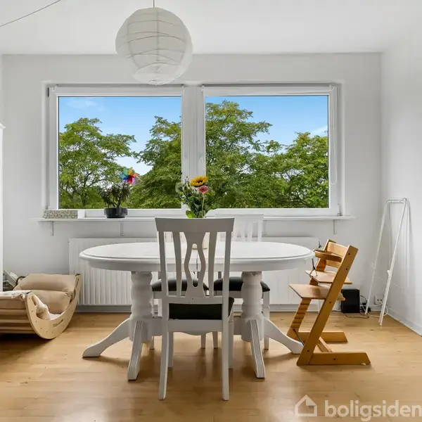 Et rundt spisebord med stole står foran et stort vindue med udsigt til grønne træer. Til venstre ses en reol og en lænestol, mens højre viser en balkon.