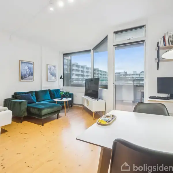 En moderne stue med en grøn sofa og vægkunst, placeret nær et stort vindue med udsigt over lejlighedsbygninger. Et tv står ved siden af, og et skrivebord med computer er til højre. En spiseplads med hvide stole er i forgrunden.