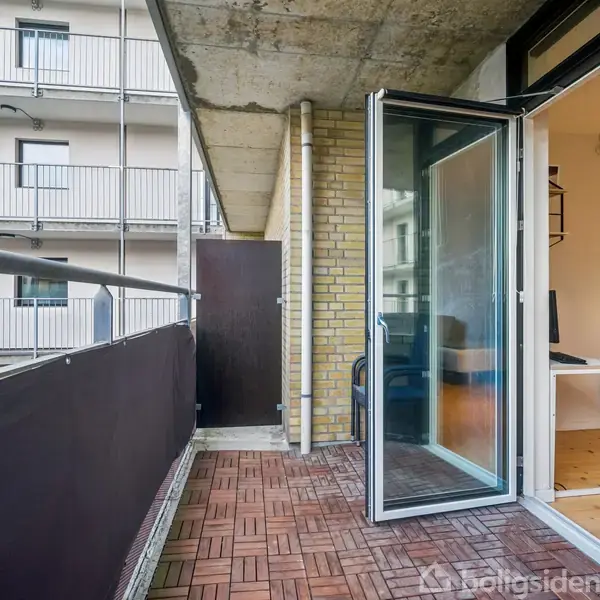 Balkon med træfliser, nogle åbne glasdøre leder til en stue med bord og stol, omgivet af en bygningsfacade med altaner.