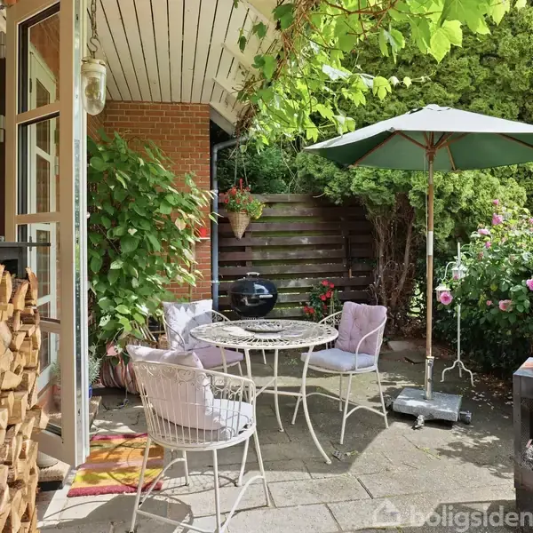 Terrasse med runde havebord og fire stole, omgivet af frodig vegetation. En parasol står midt i området, mens en stabel brænde og en lille grill skaber en hyggelig atmosfære.