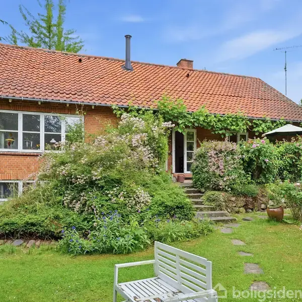 Rødt murstenshus med rødt tegltag omgivet af frodig have. Grene og buske dækker huset og flere blomster står i fuldt flor, mens en hvid bænk står i forgrunden.