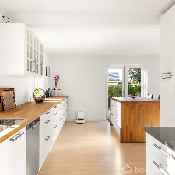 Et køkken med en stor træø omgivet af to sorte barstole. Et frugtfad står på bordet. Lys træfarvet gulv, hvidt interiør, vinduer og moderne apparater. En orkidé på skænken.