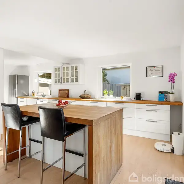 Et moderne køkken med hvide skabe og træbordplader; der står en vask, en ovn, og et stort gråt køleskab til højre. Rummet er lyst og ryddeligt.