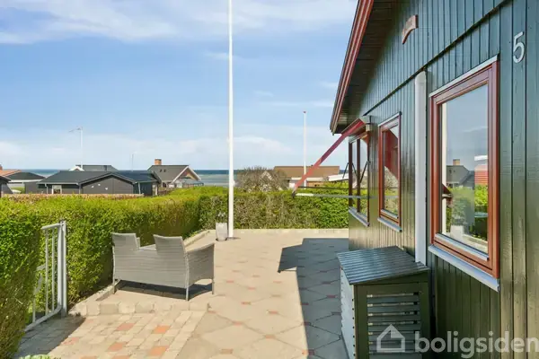 Et træhus med nummeret "5" på væggen står ved siden af en brolagt terrasse med havemøbler. Haven har klippede hække, og der er udsigt til en fjern havhorisont.