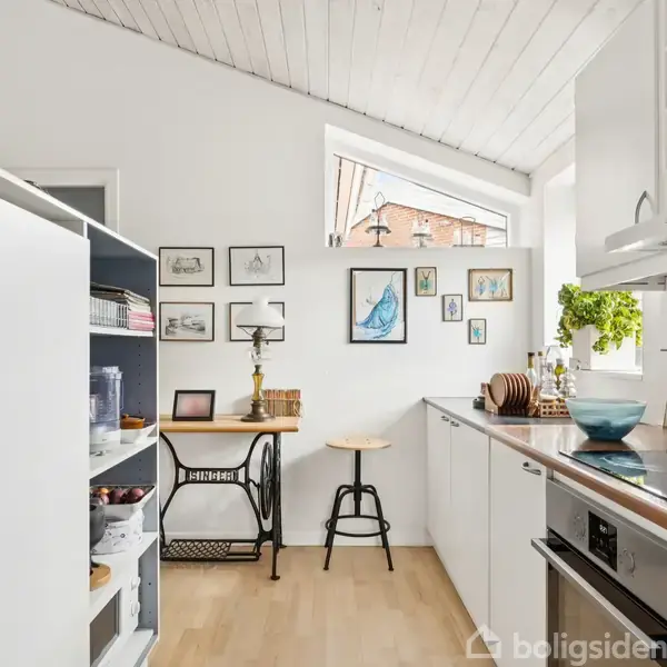 Et køkken med hvide skabe, træbordplader og enovn. En stol og en bordlampe står ved en væg dekoreret med billeder. Et lille vindue lader dagslyset komme ind.