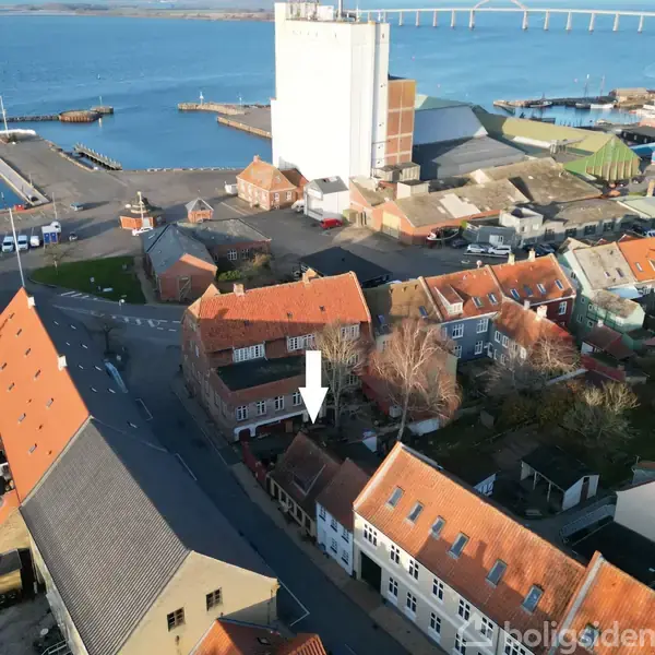 Hus med rød tag er markeret med en hvid pil. Det ligger blandt andre bygninger i et havneområde med udsigt over vandet og en bro i baggrunden.