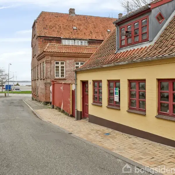En gul bygning med røde vinduer står langs en brostensbelagt gade. I baggrunden ses en større murstensbygning og en udsigt til vandet. En bil er parkeret på vejen.