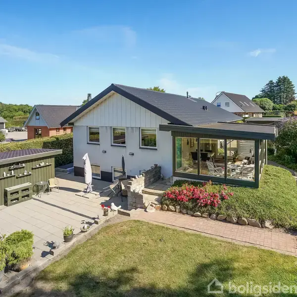 Et hvidt hus med sorte tagbjælker står i en have. Huset har en lukket veranda, omgivet af blomsterbede og en flisebelagt terrasse med havemøbler.