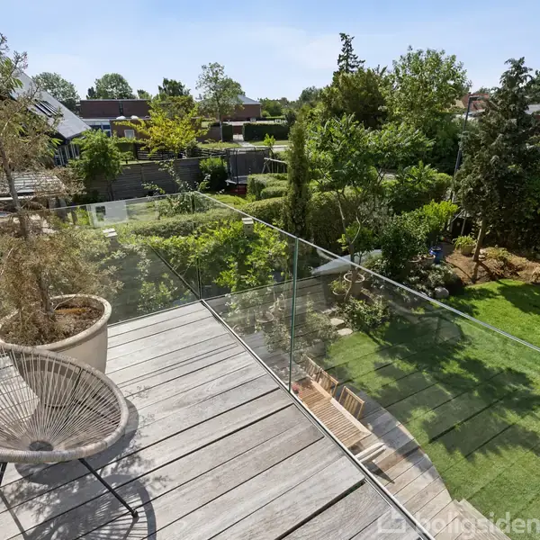 Balkon med glasrækværk, en kurvestol og en plante i en krukke; set mod en velholdt have med græsplæne og grønne træer i et villakvarter.