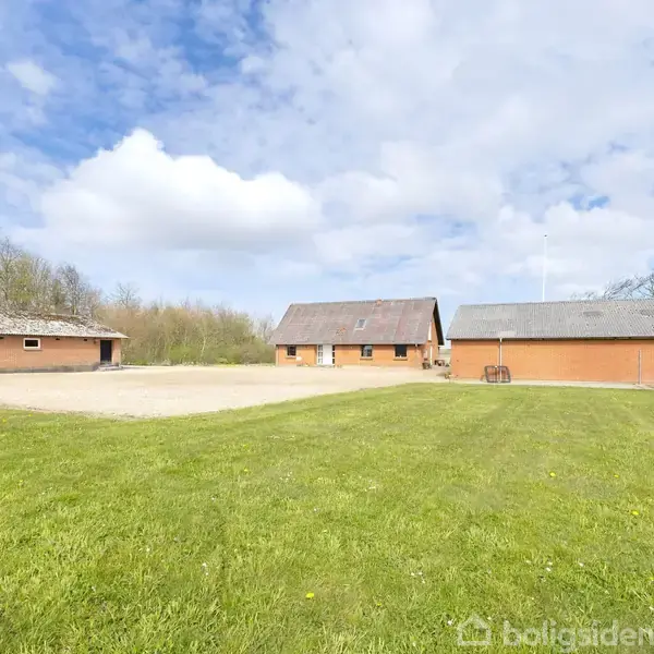 Tre røde bygninger står stille omkring en grusgårdsplads, omgivet af grønne græsarealer og træer under en blå himmel med skyer.