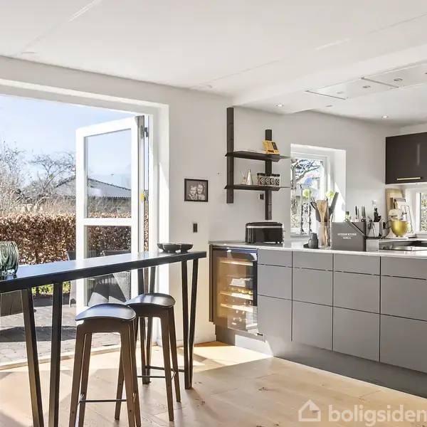 Køkkenalrum med gråt køkkenskab og sort bord med høje stole. Sollys strømmer ind gennem åbne døre til haven med hæk og buske i baggrunden.