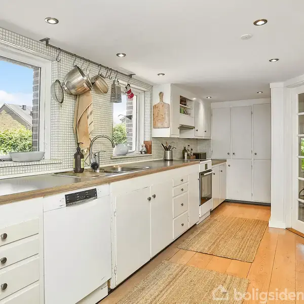 Et lyst køkken med hvide skabe og træbordplade. Krukker og gryder hænger over vasken. Bordpladerne er dekoreret med planter og redskaber. Stor dobbeltdør åbner til en udendørs udsigt.