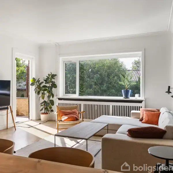 En stue med en hvid sofa står mod en blå væg, suppleret af et sofabord. Et fjernsyn står på et stativ. Store vinduer giver udsigt til en have.
