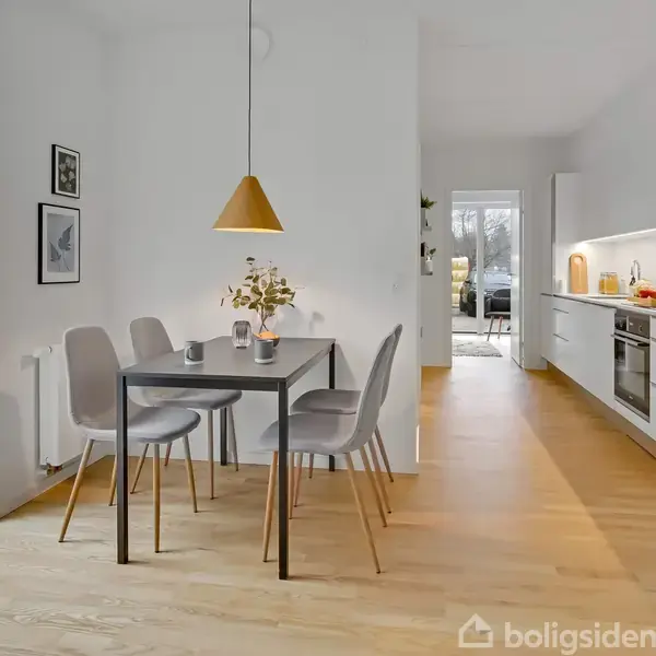 En spisestue har et bord med fire stole og en gul lampe hængende over. Køkkenet i baggrunden har hvide skabe og moderne apparater. Der er lysindfald fra store vinduer.
