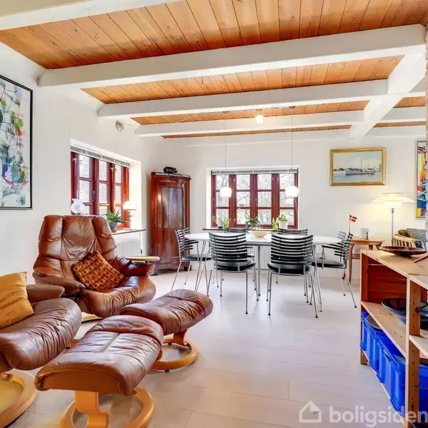 Brune lænestole står i en stue med træloft, foran vinduer og et spisebord med sorte stole. Kunstværker og lamper dekorerer rummet, der er lyst og indbydende.