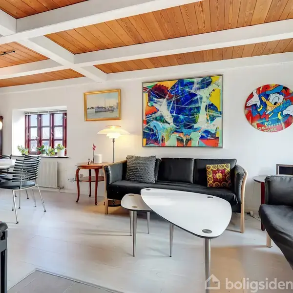 Stue med sorte sofaer og moderne sofaborde; farverige kunstværker på væggen. Træloft og hvide vægge skaber et lyst rum med spisebord i baggrunden nær et vindue.