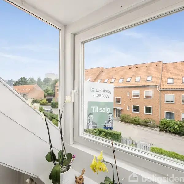 Plakat med teksten "LokaltBolig 44 88 03 03 Til salg" hænger i vindue. Udsigt til rødt tegltag, boligblokke og grønne planter udenfor. Indendørs ses orkidéer i vindueskarmen.