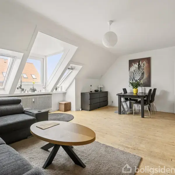 Stue med grå sofa og gule puder ved store skråvinduer. Rundt sofabord, spisebord med stole til venstre, og kommode med plante til højre i lys indretning.