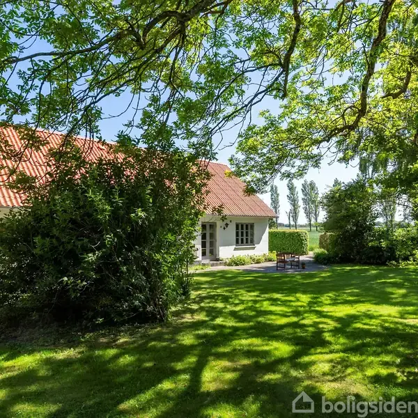Hus med rødtag og hvide vægge står stille i en frodig have med græs og træer, omgivet af grønt løv under en lys blå himmel.