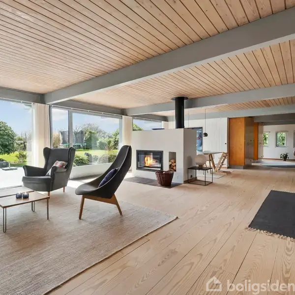Stue med moderne møbler, herunder stole og sofabord, omkring en åben pejs. Store vinduer giver udsigt til en grøn have. Lyst træloft og gulv skaber en indbydende atmosfære.