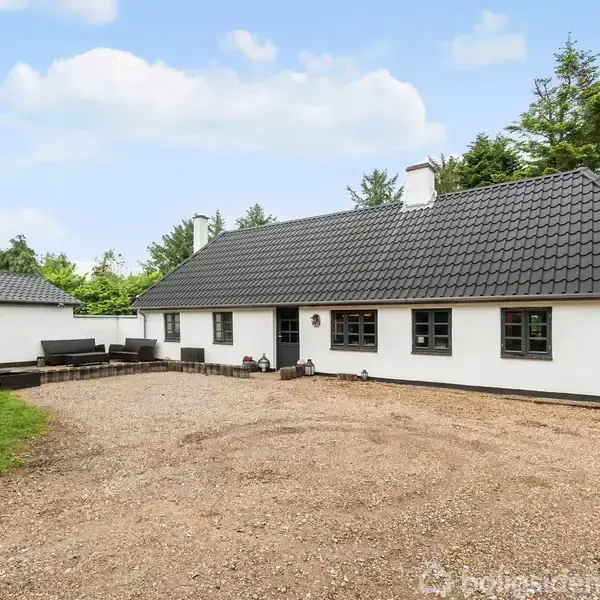Et hvidt, enkelt-etagers hus med sort tag står på en grusbelagt gårdsplads. Ved siden af huset ses en mindre bygning. Omgivet af grønne træer under en blå himmel.