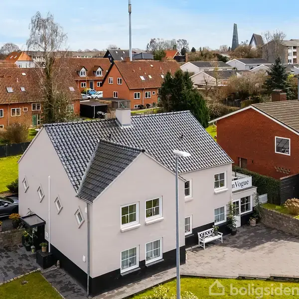 Et hvidt hus med gråt tag og flere vinduer står i en forstad med grønne områder og nabobygninger. Et skilt på huset viser teksten "inVogue".