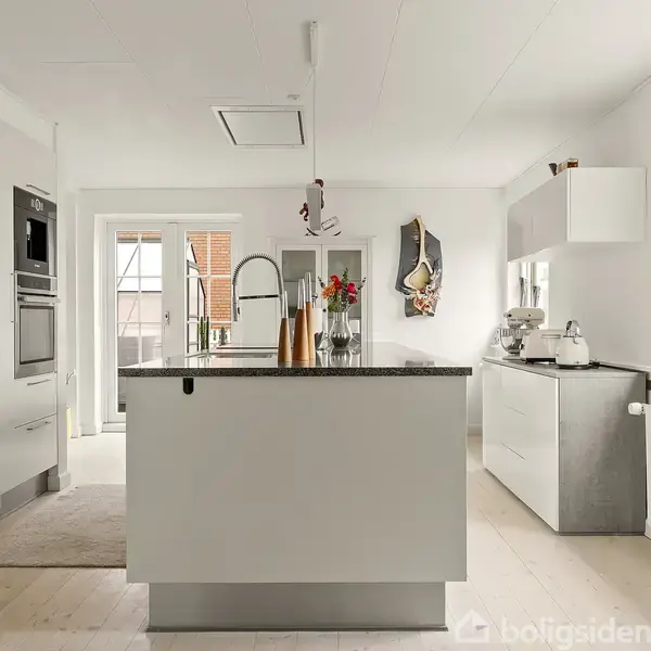 En moderne køkkenø med en vask og blandingsbatteri dominerer rummet, omgivet af hvide skabe og apparater. Et vindue til højre giver naturligt lys, mens en kaffemaskine står på en skænk.