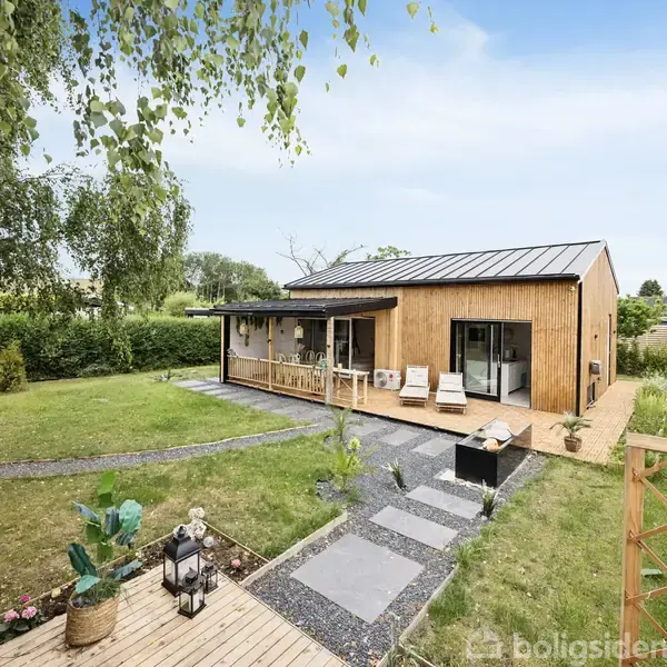 Moderne træhus med sort tag står i frodig have. To liggestole er placeret på terrasse; stier leder gennem det grønne græs. Blå himmel over rolige omgivelser.