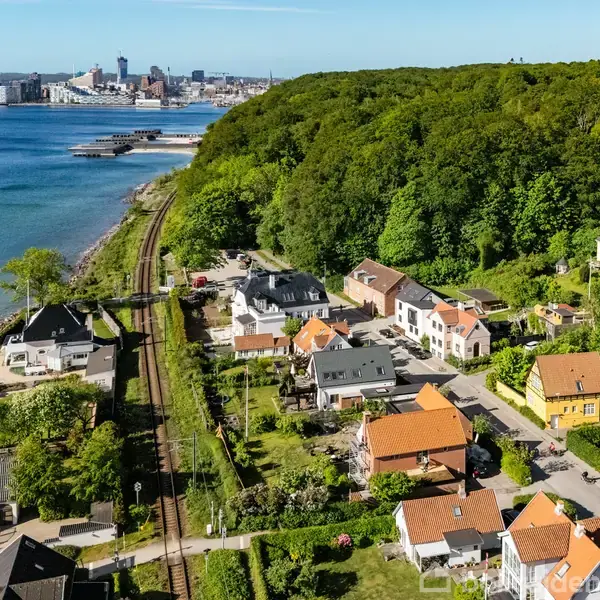 Række af huse langs en jernbane i frodig grøn landskab. Havet ligger til venstre, mens skoven strækker sig i baggrunden. Byens skyline ses i horisonten.
