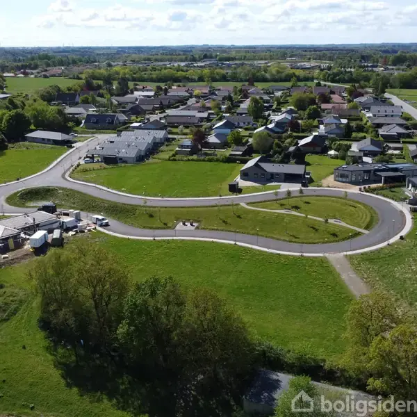 Nybyggeri er omgivet af grønne marker i en forstad. Veje er snoede med flere byggegrunde, nogle med begyndende konstruktion. Boliger og træer er synlige i baggrunden.
