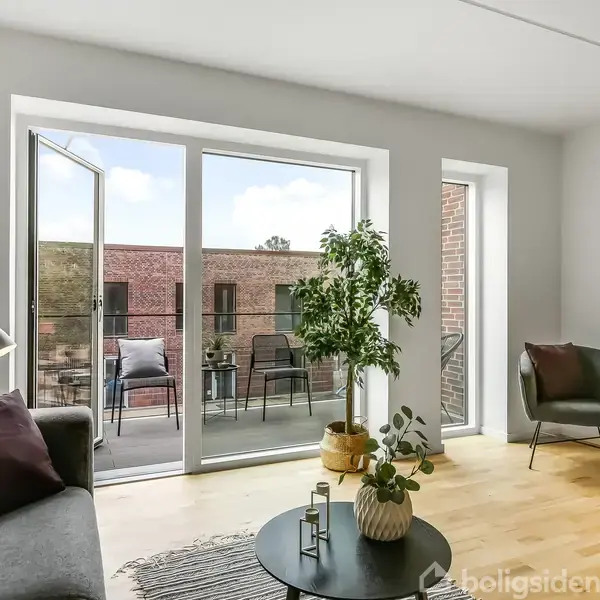 Stue med en sofagruppe med grå sofa og lænestol, plante på et lille bord, åbner ud til en altan med stole. Lysindfald fra store glasdøre.