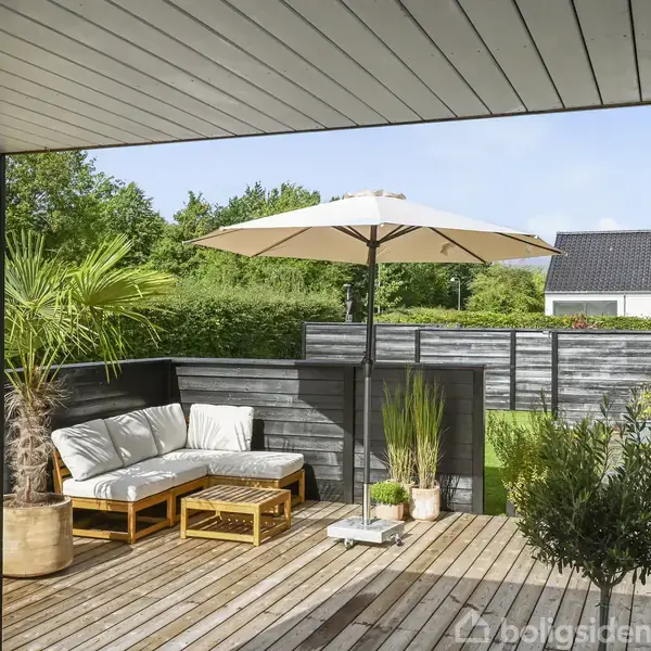Terrasse med træbrædder og havemøbler inklusiv sofa og bord, står under en stor parasol. Planter flankerer området, og høje buske omkranser terrassen, hvilket skaber en hyggelig atmosfære.