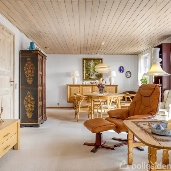 Stue med brunt læderlænestol og sofabord i forgrunden, fladskærms-TV til venstre. Middagsbord med stole og skænk i baggrunden. Lyse vægge og trægulv fuldender rummet.