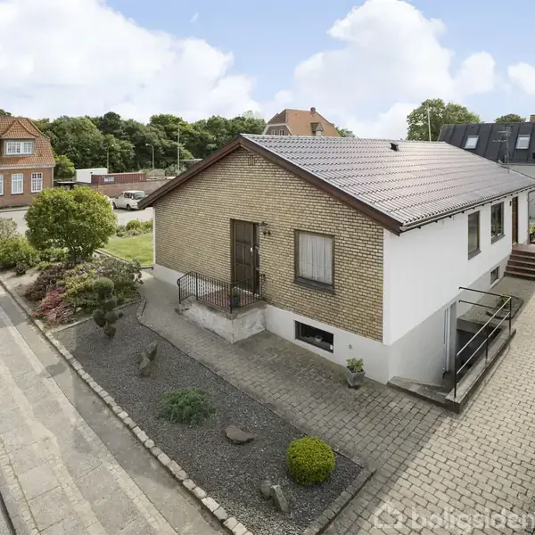 Et parcelhus i mursten med skråt tag står på en brolagt vej. Haven er anlagt med buske og sten. Flere huse og et grønt område ses i baggrunden.