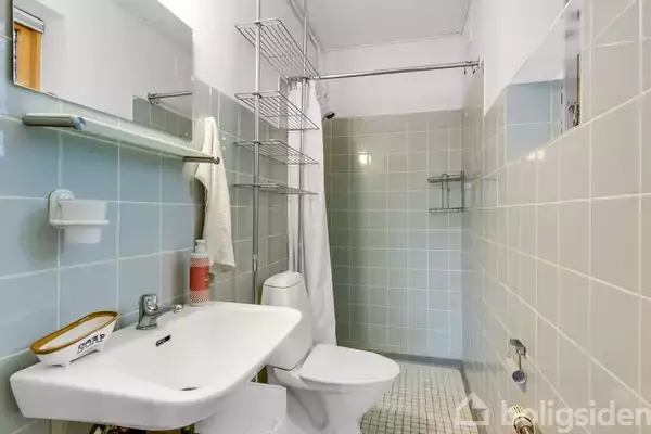 Håndvask med sæbeskål til venstre, toilet ved siden af, og en bruseniche med hvidt forhæng. Fliser dekorerer vægge og gulv. En håndklæderække hænger på væggen.