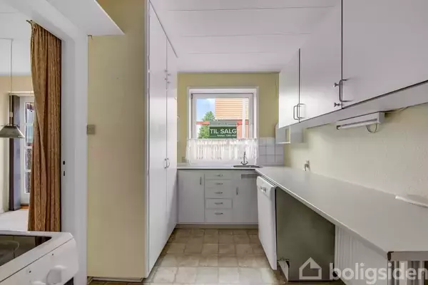 Et lyst køkken med hvide skabe og skuffer, køkkenvask under et vindue. Udenfor er et skilt med teksten “TIL SALG”. En tilstødende stue ses til venstre.