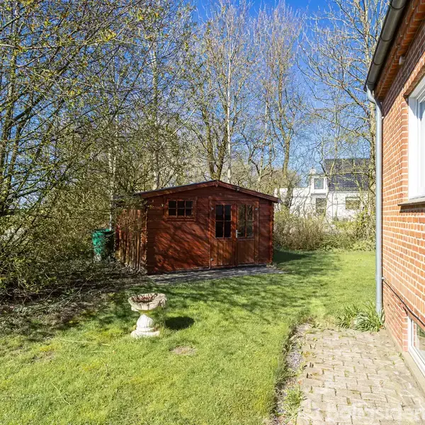 Rødt murstenshus med vindue til højre, træskur i baggrunden, græsplæne og brostenssti omgivet af træer, under en blå himmel. En lille piedestal står på græsset.