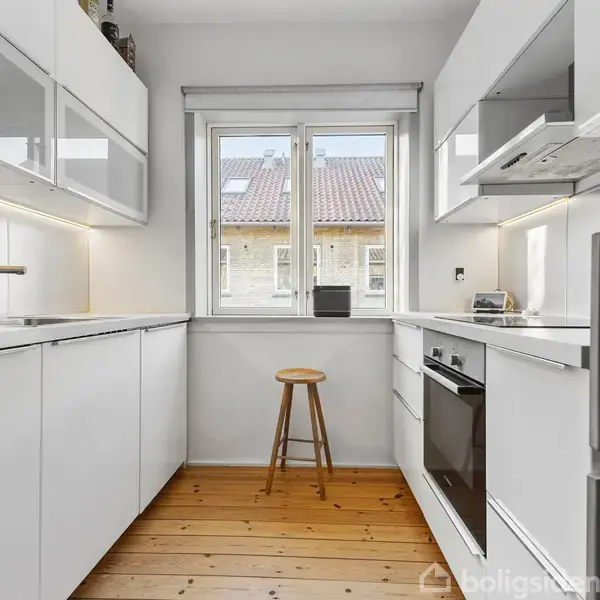 Et moderne køkken med hvide skabe og apparater. En skammel står foran et vindue, som lader naturligt lys ind. Trægulv og en ovn er synlige i rummet.