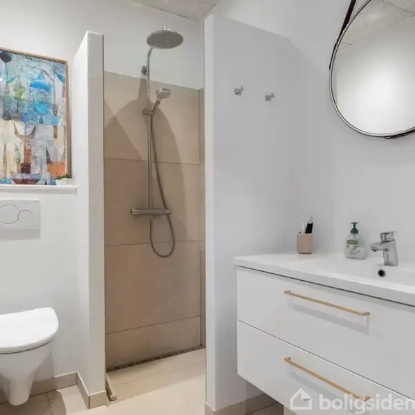 Et moderne badeværelse indeholder et væghængt toilet og bruser med regnvandsbrusehoved. Til højre er en hvid håndvask med to skuffer. Et rundt spejl hænger over vasken, og et farverigt maleri hænger ved toilettet.