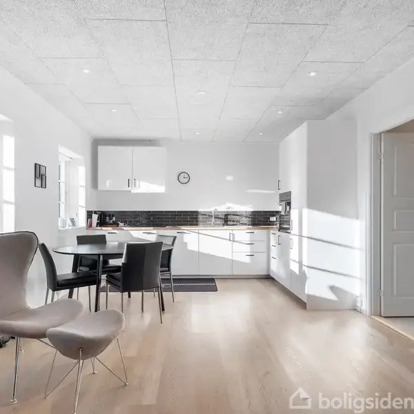 En grå lænestol står i midten af et moderne køkkenalrum med hvidmalede vægge. Køkkenet har hvide skabe og en sort bagvæg. Sollys kommer ind gennem store vinduer.