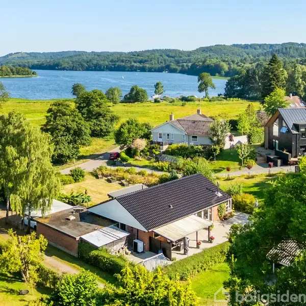 Hus med mørkt tag ligger i et grønt landskab, omgivet af træer og marker. I baggrunden ses en stor sø og skovklædte bakker under en blå himmel.