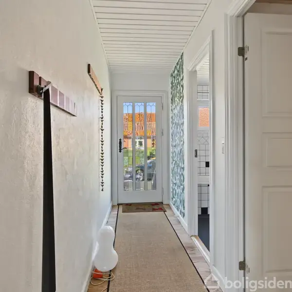 En lys gang fører mod en glasdør, hvor et stribet tæppe ligger på gulvet. Til højre er der en åben dør til et flisebelagt badeværelse.