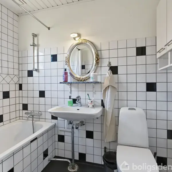 Et badeværelse med hvidt flisebelagt væg med sorte detaljer. En oval spejl hænger over en håndvask med hylde. Badekar til venstre, toilet til højre, og håndklæde hænger ved siden af spejlet.