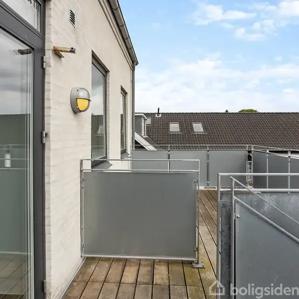 Altan med glasværn og trædækning, set fra siden. Bygningen er hvid, med glasdør og væglampe. I baggrunden ses tagene af nærliggende strukturer under en blå himmel.