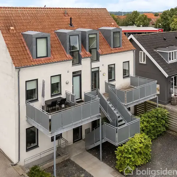 En hvid bygning med rødt tegltag har to altaner og ydre trapper i metal. Det ligger i et boligområde med en bred vej ved siden af og grønne træer i baggrunden.