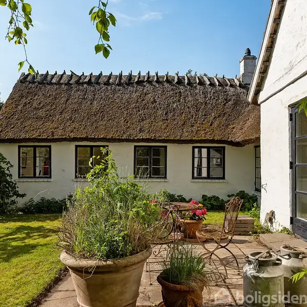 Stråtækt hus med hvide mure og sorte vinduer, omgivet af en frodig have med græsplæne og krukker. Et bord og stole står på en terrasse i solskin.