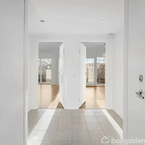 Dobbeltdør åbner ud til stue med trægulve, indrammet af hvide vægge. Store vinduer i baggrunden slipper lys ind og giver udsigt til have.