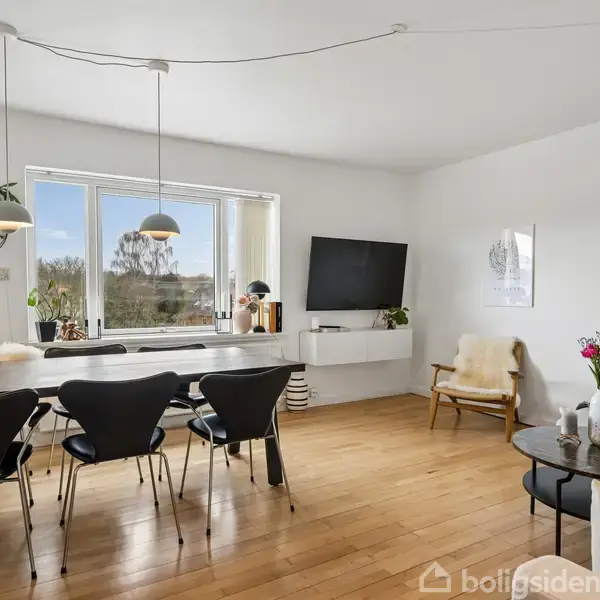 En lys stue med et spisebord og sorte stole står centralt. Til højre ses en TV på væggen og en stol med et tæppe. Store vinduer giver udsigt til naturen udenfor. En sofa og sofabord står længere væk.